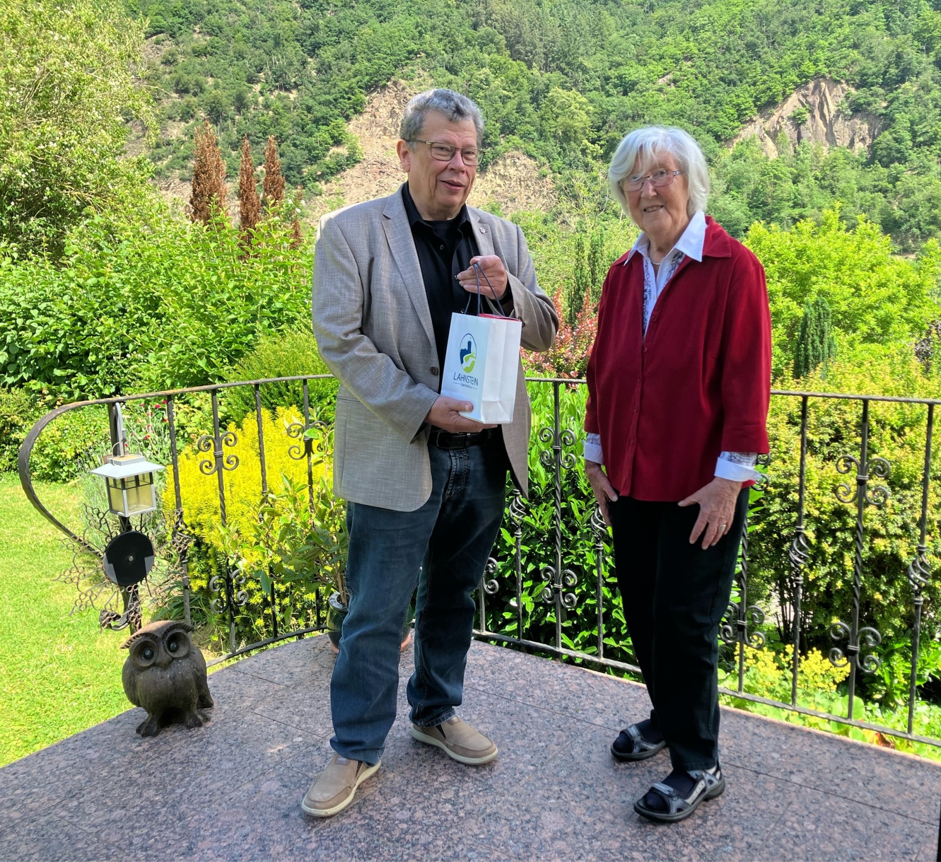 Eine ältere Frau und ein Mann stehen gemeinsam auf einer Terrasse mit schmiedeeisernem Geländer vor einer grünen, bewaldeten Hügellandschaft. Der Mann hält eine kleine Geschenktüte in der Hand und wendet sich lächelnd der Frau zu, die ebenfalls freundlich in die Kamera blickt. Neben ihnen steht eine dekorative Eulenfigur, im Hintergrund erstreckt sich eine sonnige Naturkulisse.