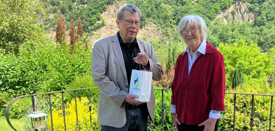 Alke Stahlschmidt feierte ihren 90. Geburtstag in Lahnstein Eine ältere Frau und ein Mann stehen gemeinsam auf einer Terrasse mit schmiedeeisernem Geländer vor einer grünen, bewaldeten Hügellandschaft. Der Mann hält eine kleine Geschenktüte in der Hand und wendet sich lächelnd der Frau zu, die ebenfalls freundlich in die Kamera blickt. Neben ihnen steht eine dekorative Eulenfigur, im Hintergrund erstreckt sich eine sonnige Naturkulisse.