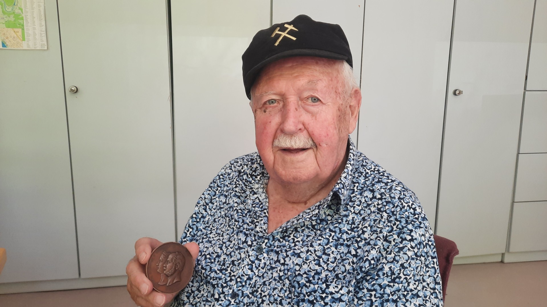 Älterer Mann sitzt in einem Raum vor Schränken und hält eine historische Medaille in der Hand; er trägt ein gemustertes Hemd und eine dunkle Kappe und blickt in die Kamera.