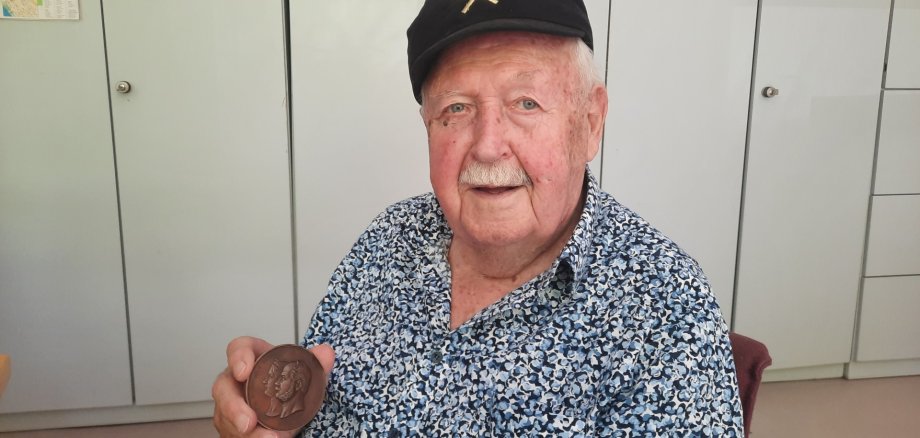 Älterer Mann sitzt in einem Raum vor Schränken und hält eine historische Medaille in der Hand; er trägt ein gemustertes Hemd und eine dunkle Kappe und blickt in die Kamera.
