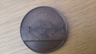 Alte Kupfermedaille liegt auf einem hellen hölzernen Hintergrund. Die Medaille zeigt auf ihrer Vorderseite eine Brücke und dahinter die Festung Ehrenbreitstein.