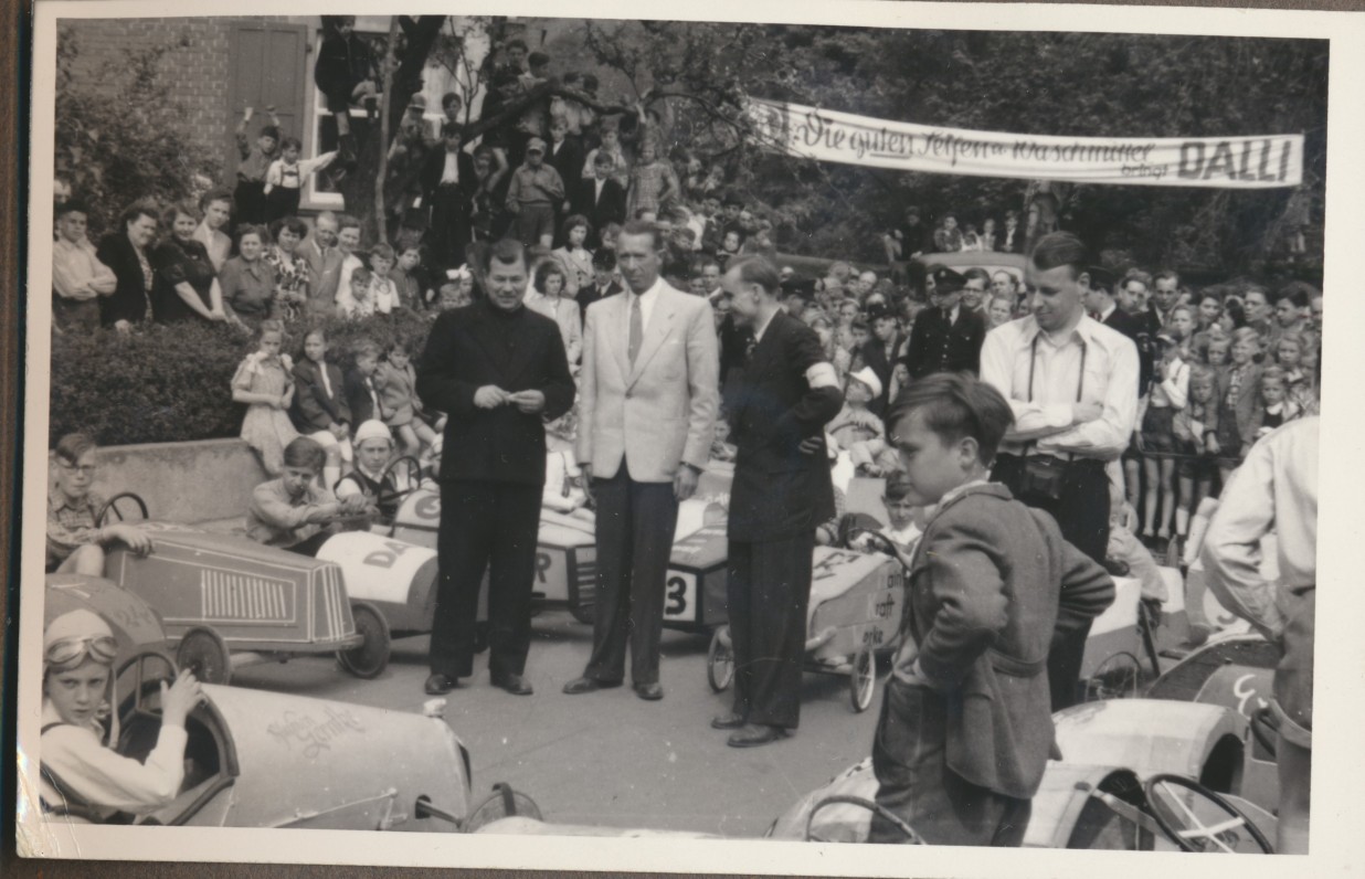 Eine historische Schwarz-Weiß-Fotografie eines Seifenkistenrennens in Oberlahnstein um 1950. Im Vordergrund und Hintergrund stehen zahlreiche selbstgebaute Seifenkisten mit jungen Fahrern in Rennhauben und Fliegerbrillen. Im Zentrum stehen drei erwachsene Männer in Anzügen inmitten der Menge; ganz links ein Geistlicher im Talar. Dahinter drängen sich zahlreiche Zuschauer an einem Hang. Im Hintergrund hängt ein langes Werbebanner mit der Aufschrift „Die guten Seifen u. Waschmittel bringt DALLI“.