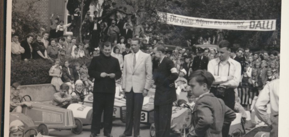 Eine historische Schwarz-Weiß-Fotografie eines Seifenkistenrennens in Oberlahnstein um 1950. Im Vordergrund und Hintergrund stehen zahlreiche selbstgebaute Seifenkisten mit jungen Fahrern in Rennhauben und Fliegerbrillen. Im Zentrum stehen drei erwachsene Männer in Anzügen inmitten der Menge; ganz links ein Geistlicher im Talar. Dahinter drängen sich zahlreiche Zuschauer an einem Hang. Im Hintergrund hängt ein langes Werbebanner mit der Aufschrift „Die guten Seifen u. Waschmittel bringt DALLI“.