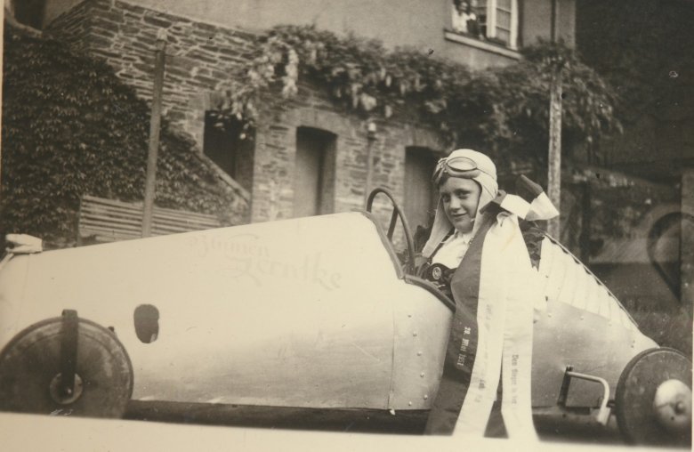 Eine historische Schwarz-Weiß-Nahaufnahme eines jungen Rennfahrers, der stolz in seiner aerodynamisch gebauten, hellen Seifenkiste sitzt. Das Fahrzeug trägt die Aufschrift „Blumen Gerntke“. Der Junge trägt eine Fliegerhaube mit Rennbrille auf dem Kopf und eine breite Siegerschleife über der Schulter, auf der das Datum „20. Mai 1951“ gedruckt ist. Im Hintergrund ist die Fassade eines älteren, mit Efeu bewachsenen Gebäudes zu sehen.