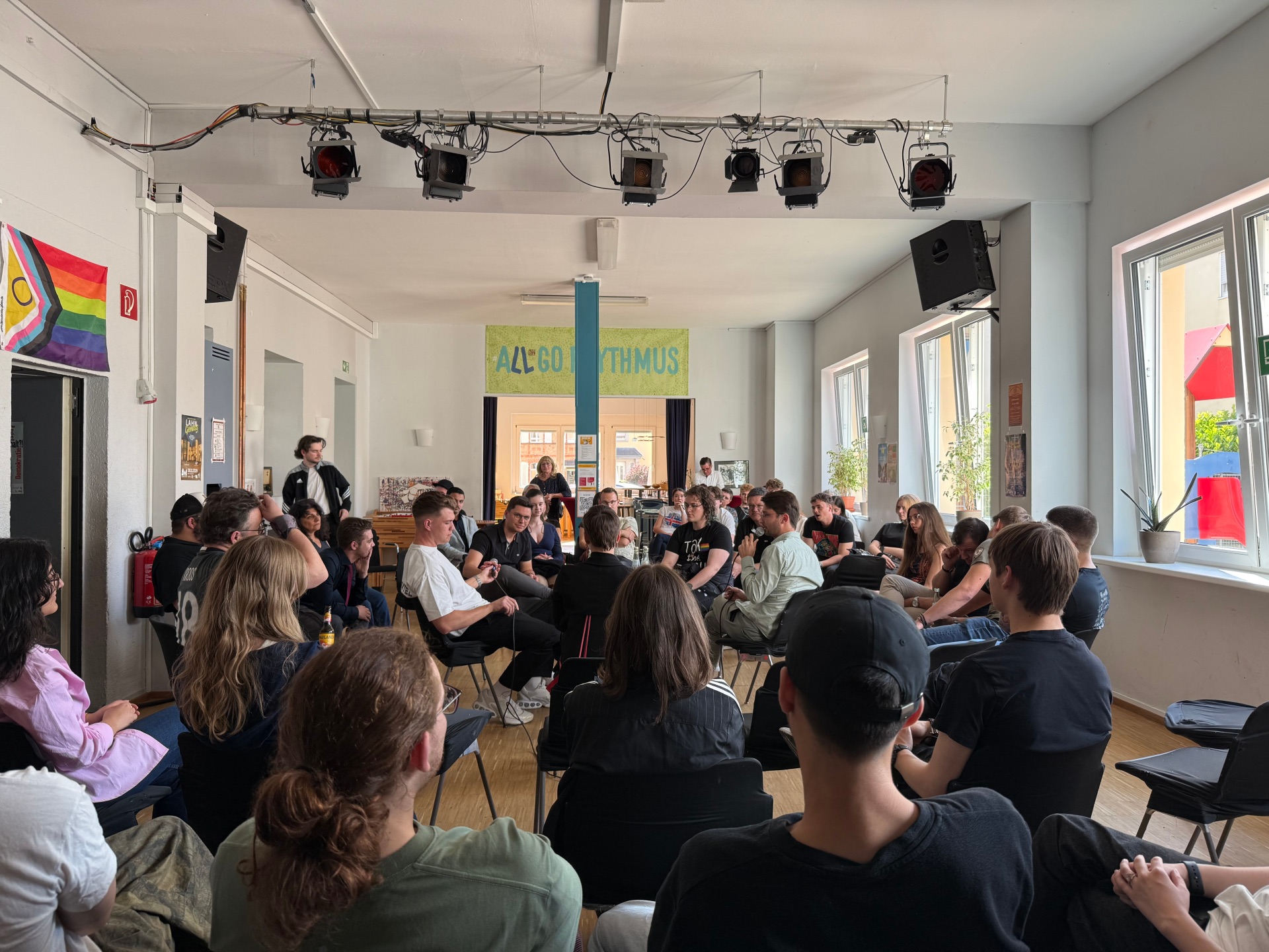 Ein heller Raum mit vielen jungen Menschen, die in einem großen Stuhlkreis sitzen und diskutieren. Einige melden sich oder sprechen, während andere zuhören. An der Wand hängt eine Regenbogenflagge, und im Hintergrund ist ein Schild mit der Aufschrift „All Good Rhythmus“ zu sehen. Die Atmosphäre wirkt offen und engagiert.