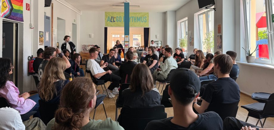 Junge Menschen diskutierten im Jugendkulturzentrum mit Politikern über die Zukunft Ein heller Raum mit vielen jungen Menschen, die in einem großen Stuhlkreis sitzen und diskutieren. Einige melden sich oder sprechen, während andere zuhören. An der Wand hängt eine Regenbogenflagge, und im Hintergrund ist ein Schild mit der Aufschrift „All Good Rhythmus“ zu sehen. Die Atmosphäre wirkt offen und engagiert.
