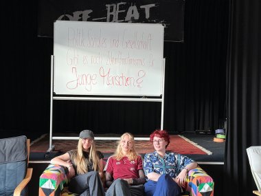 Junge Menschen diskutierten im Jugendkulturzentrum mit Politikern über die Zukunft Drei junge Menschen sitzen entspannt auf einem bunten Sofa vor einer Bühne. Hinter ihnen steht ein Whiteboard mit der handgeschriebenen Frage „Politik, Soziales und Gesellschaft: Gibt es noch Zukunftsoptimismus für junge Menschen?“. Die drei schauen in die Kamera und wirken freundlich und aufmerksam.