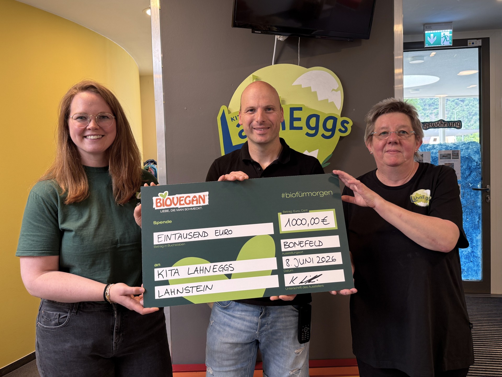 Drei Personen stehen im Eingangsbereich der kommunalen Kita LahnEggs in Lahnstein und präsentieren einen symbolischen Förderscheck über 1.000 Euro aus dem Projekt „#biofürmorgen“ von BIOVEGAN. Im Hintergrund ist das Logo der Kita LahnEggs zu sehen.