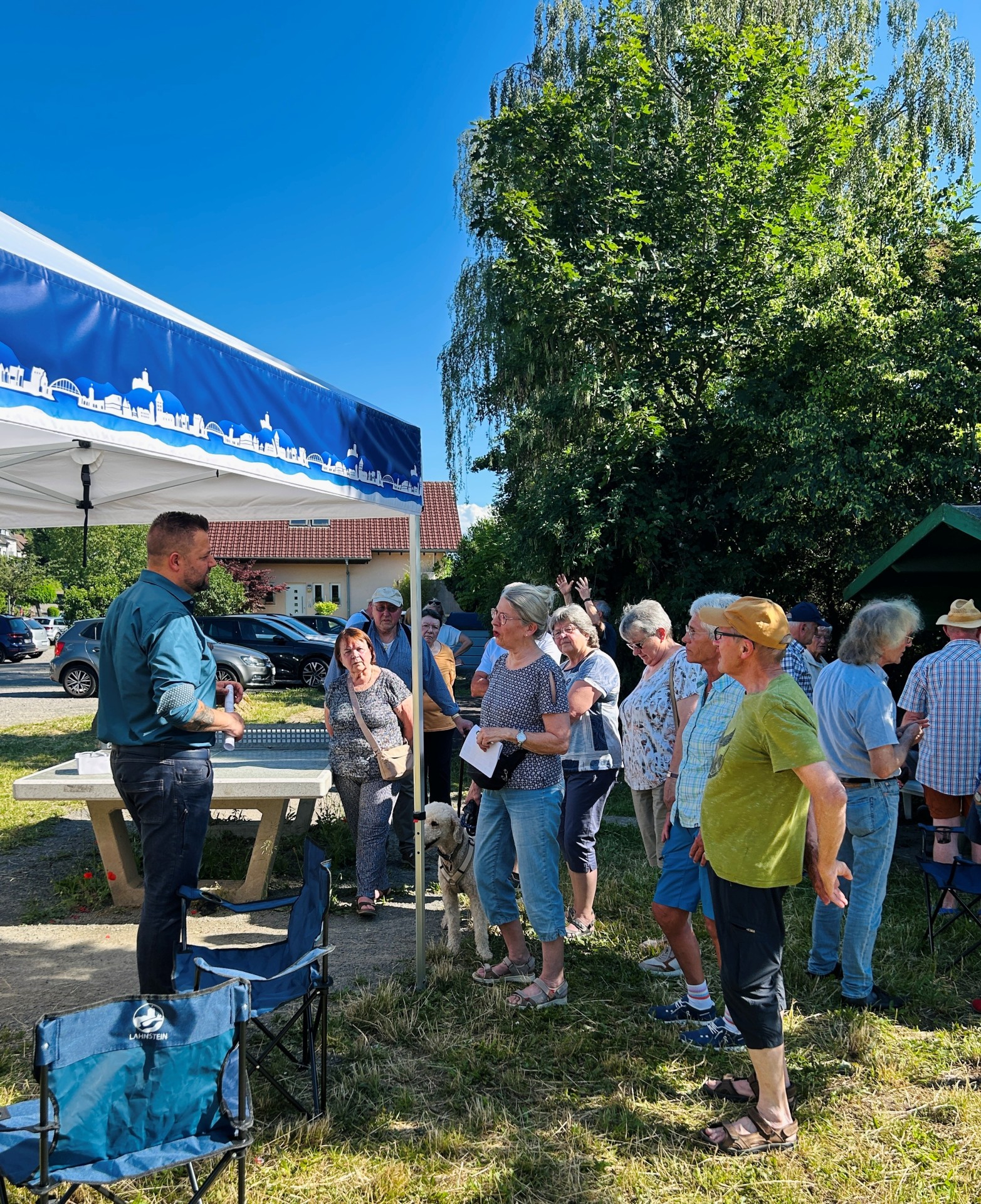 Das Foto zeigt eine sommerliche Außenaufnahme einer Bürgerversammlung oder Infoveranstaltung im Freien. Auf einer Wiese mit einer Tischtennisplatte im Hintergrund und dichten grünen Bäumen hat sich eine Gruppe von etwa einem Dutzend Menschen mittleren bis reiferen Alters versammelt. Im Zentrum der Gruppe steht ein Mann im dunkelgrünen Hemd und Jeans, der zu den Zuhörern spricht und mit einer Frau in vorderster Front interagiert. Die Frau hat graues Haar, trägt ein gemustertes T-Shirt und hellblaue Jeans und hat einen hellbeigen Pudel an der Leine. Die anderen Anwesenden lauschen aufmerksam, einige halten Handzettel. Links im Vordergrund ist der Rand eines Pavillons mit einer blauen Blende zu sehen, die eine weiße Skyline-Silhouette zeigt. Zwei blaue Campingstühle mit der Aufschrift „Lahnstein“ stehen ebenfalls links. Die Stimmung wirkt entspannt und informell. Das Licht ist helles Tageslicht bei klarem blauem Himmel.