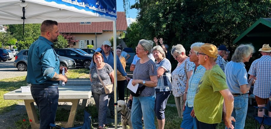 „Lennart Live“ auf dem Allerheiligenberg Das Foto zeigt eine sommerliche Außenaufnahme einer Bürgerversammlung oder Infoveranstaltung im Freien. Auf einer Wiese mit einer Tischtennisplatte im Hintergrund und dichten grünen Bäumen hat sich eine Gruppe von etwa einem Dutzend Menschen mittleren bis reiferen Alters versammelt. Im Zentrum der Gruppe steht ein Mann im dunkelgrünen Hemd und Jeans, der zu den Zuhörern spricht und mit einer Frau in vorderster Front interagiert. Die Frau hat graues Haar, trägt ein gemustertes T-Shirt und hellblaue Jeans und hat einen hellbeigen Pudel an der Leine. Die anderen Anwesenden lauschen aufmerksam, einige halten Handzettel. Links im Vordergrund ist der Rand eines Pavillons mit einer blauen Blende zu sehen, die eine weiße Skyline-Silhouette zeigt. Zwei blaue Campingstühle mit der Aufschrift „Lahnstein“ stehen ebenfalls links. Die Stimmung wirkt entspannt und informell. Das Licht ist helles Tageslicht bei klarem blauem Himmel.