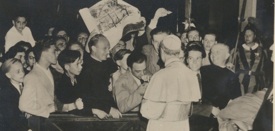 Historische Schwarz-Weiß-Aufnahme in einer Kirche: Eine Gruppe überwiegend junger Menschen drängt sich um einen Geistlichen und einen hochrangigen Kirchenvertreter in liturgischer Kleidung. Die Jugendlichen blicken aufmerksam auf das Geschehen, einige halten ein großes Geschenk oder Plakat in die Höhe. Die Szene vermittelt die feierliche und zugleich herzliche Atmosphäre einer kirchlichen Begegnung in den 1950er-Jahren.