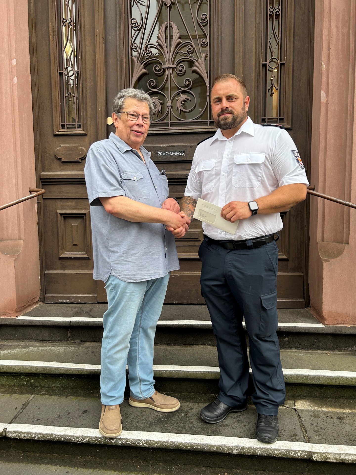 Ein Foto von zwei Männern, die sich vor einer großen, dunklen Holztür mit Metallbeschlägen und Glaseinsätzen die Hände schütteln. Der linke Mann ist älter, trägt eine Brille, ein hellblaues Kurzarmhemd, Jeans und Slipper. Der rechte Mann ist jünger, kräftig gebaut, trägt eine weiße Dienstuniform der Feuerwehr mit einem Abzeichen auf dem Ärmel, eine Smartwatch und hält einen Briefumschlag in seiner linken Hand. Sie stehen auf Steinstufen.