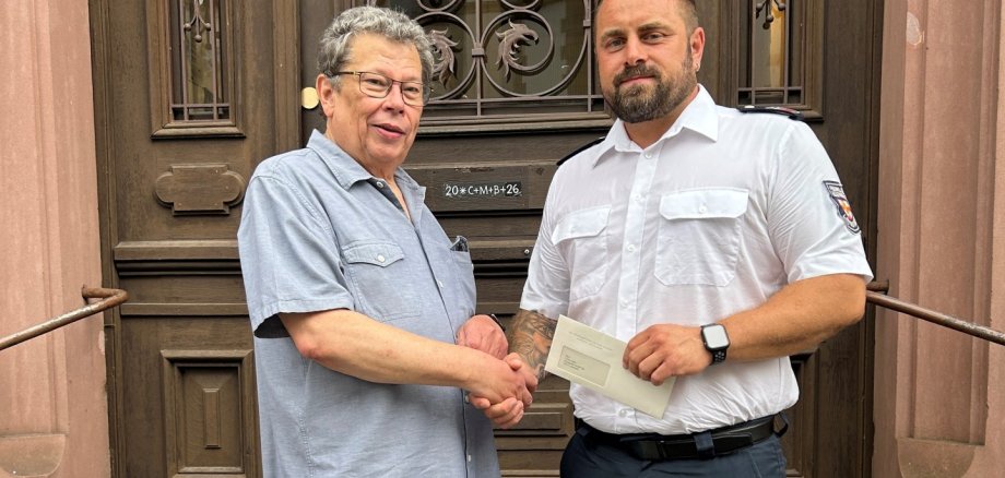 Ein Foto von zwei Männern, die sich vor einer großen, dunklen Holztür mit Metallbeschlägen und Glaseinsätzen die Hände schütteln. Der linke Mann ist älter, trägt eine Brille, ein hellblaues Kurzarmhemd, Jeans und Slipper. Der rechte Mann ist jünger, kräftig gebaut, trägt eine weiße Dienstuniform der Feuerwehr mit einem Abzeichen auf dem Ärmel, eine Smartwatch und hält einen Briefumschlag in seiner linken Hand. Sie stehen auf Steinstufen.