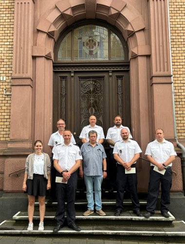 Gruppenfoto von sieben Personen, die auf den Stufen vor einem Gebäude mit Sandsteinfassade und einer kunstvollen Eingangstür stehen. Der Mann im grauen Hemd und Jeans aus dem ersten Bild steht im Zentrum. Fünf Männer in weißen Feuerwehruniformen und eine Frau in ziviler Kleidung umgeben ihn. Mehrere Feuerwehrmänner halten Briefumschläge. Ein geschwungenes Portal mit einem Wappen aus buntem Glas befindet sich über der Tür.