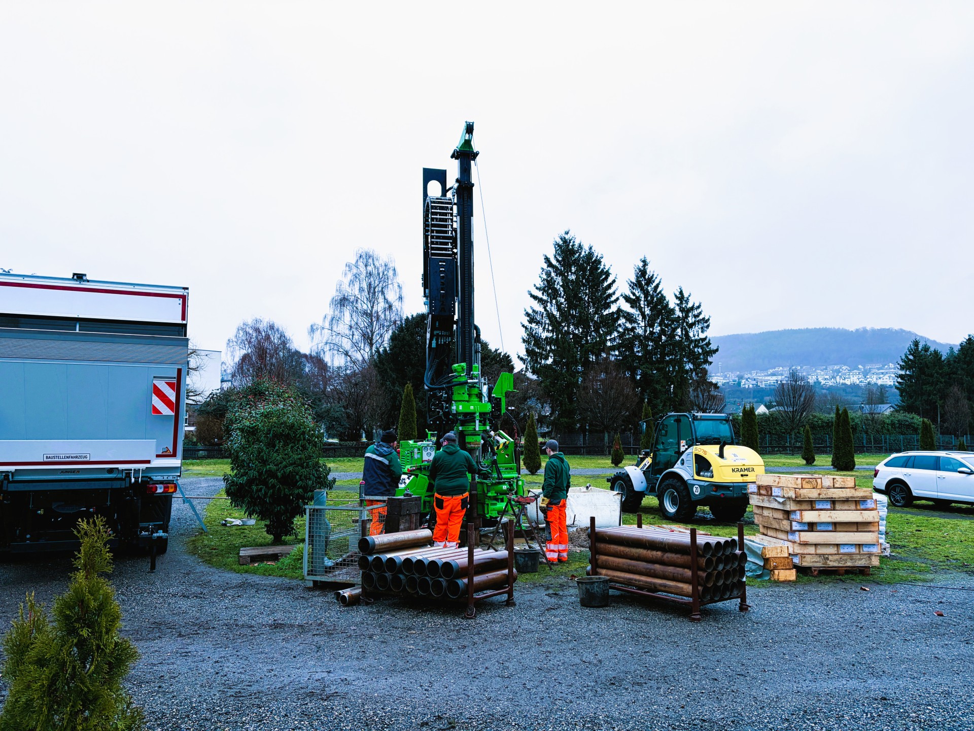 Ein Gerät für Kernbohrungen auf grüner Wiese, vor dem drei Menschen in Warnkleidung stehen, sowie die Rückseite eines LKW (links), ein PKW (rechts) und ein Baustellenfahrzeug (rechts). Dahinter sind grüne Bäume erkennbar. 