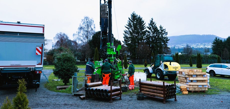 Ein Gerät für Kernbohrungen auf grüner Wiese, vor dem drei Menschen in Warnkleidung stehen, sowie die Rückseite eines LKW (links), ein PKW (rechts) und ein Baustellenfahrzeug (rechts). Dahinter sind grüne Bäume erkennbar. 