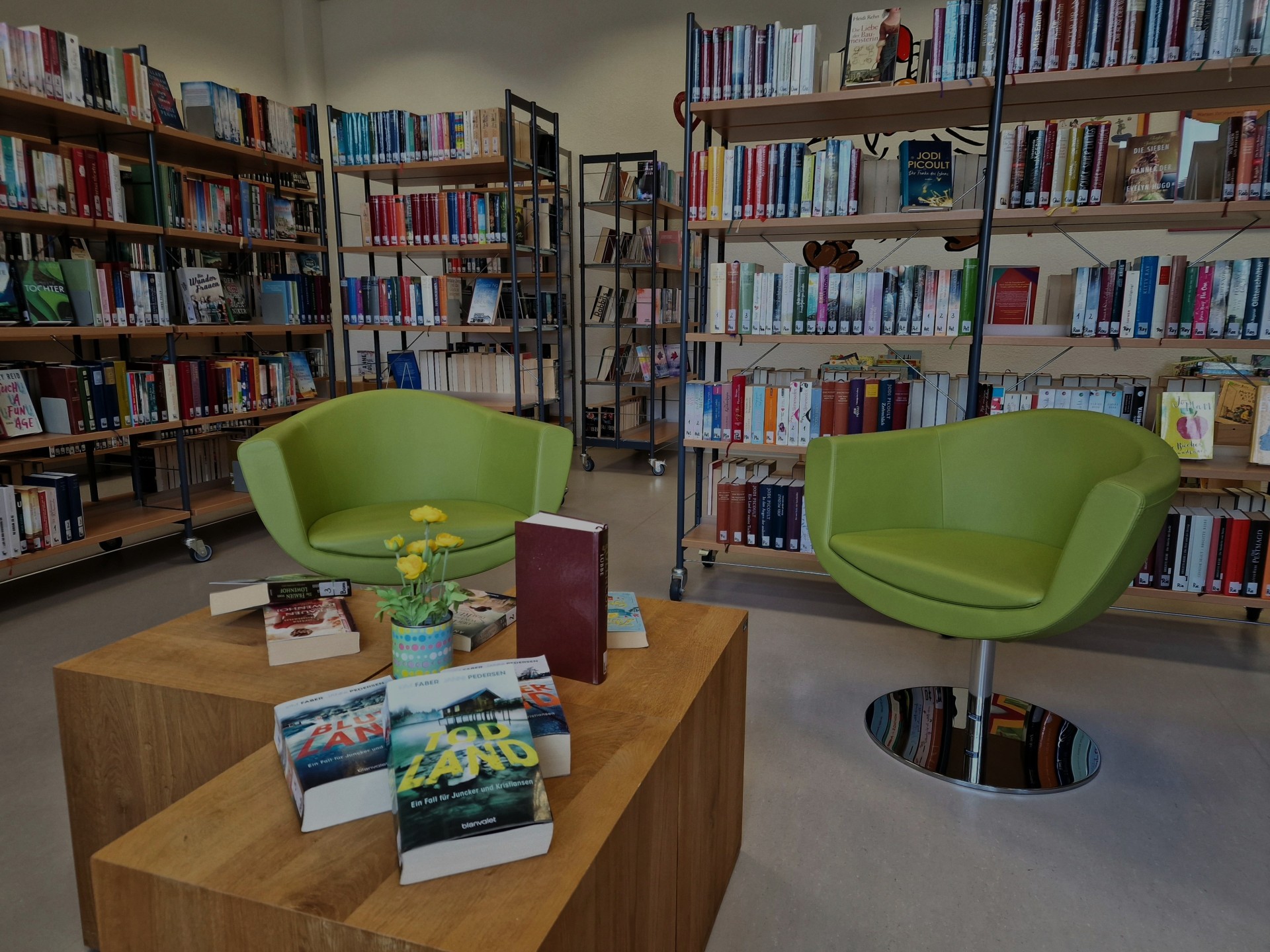 Zwei grüne Sessel stehen in der Stadtbücherei Lahnstein an einem kleinem Holztisch, auf dem sich Bücher und Blumen befinden. Im Hintergrund sind viele volle Bücherregale zu sehen.