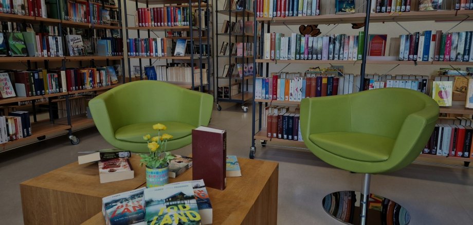 Zwei grüne Sessel stehen in der Stadtbücherei Lahnstein an einem kleinem Holztisch, auf dem sich Bücher und Blumen befinden. Im Hintergrund sind viele volle Bücherregale zu sehen.