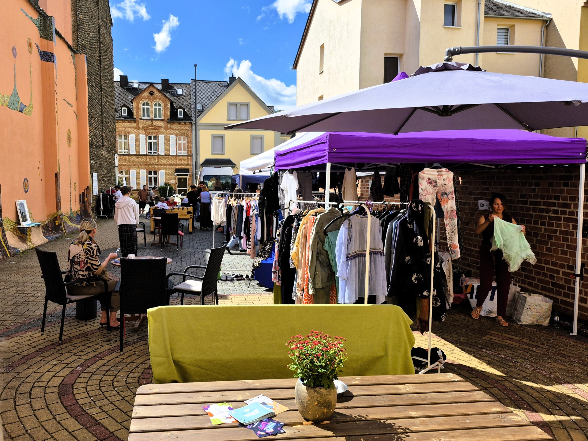 Sonniger Innenhof des Jugendkulturzentrums Lahnstein während einer Klamottenbörse mit Kleiderständern unter einem lila Pavillon, Sitzgelegenheiten an Holztischen und Menschen, die zwischen bunt gestalteten Hauswänden stöbern.