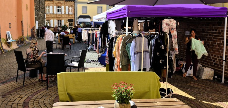 Sonniger Innenhof des Jugendkulturzentrums Lahnstein während einer Klamottenbörse mit Kleiderständern unter einem lila Pavillon, Sitzgelegenheiten an Holztischen und Menschen, die zwischen bunt gestalteten Hauswänden stöbern.