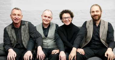 Drei Männer und eine Frau sitzen in einer Reihe nebeneinander und schauen in die Kamera.