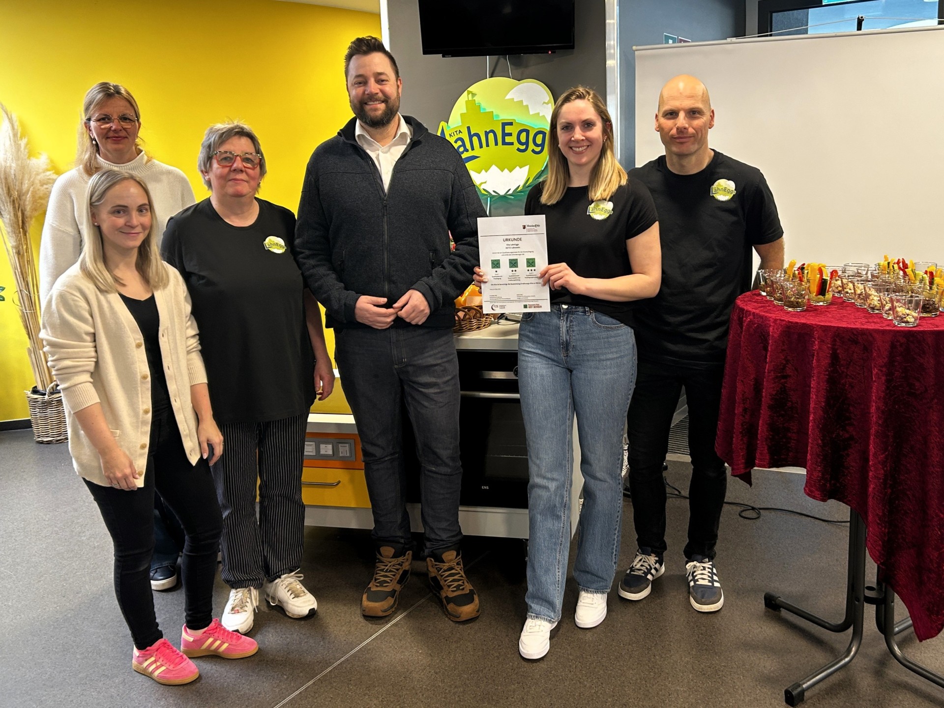 Mehrere Personen stehen nebeneinander. In der Mitte wird die Urkunde für die Auszeichnung als 3-Sterne-Ernährungskita präsentiert. Im Hintergrund ist das Logo der Kita zu sehen, rechts steht ein Tisch mit kleinen Gläsern und vorbereiteten Snacks für die Feier.
