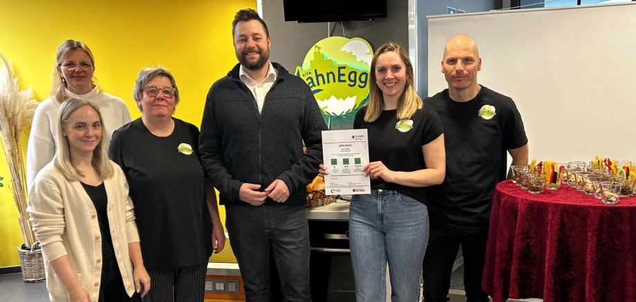 Drei Sterne für die „LahnEggs“ Mehrere Personen stehen nebeneinander. In der Mitte wird die Urkunde für die Auszeichnung als 3-Sterne-Ernährungskita präsentiert. Im Hintergrund ist das Logo der Kita zu sehen, rechts steht ein Tisch mit kleinen Gläsern und vorbereiteten Snacks für die Feier.