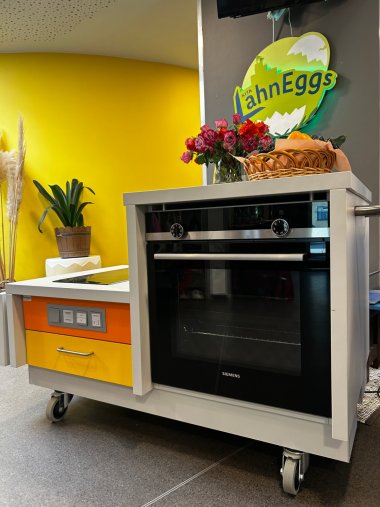 Drei Sterne für die „LahnEggs“ Eine mobile Kochstation mit Backofen steht im Eingangsbereich der Kita LahnEggs. Darüber hängt das Logo der Einrichtung. Auf der Arbeitsfläche befinden sich Blumen und ein Korb mit Obst und Gemüse.