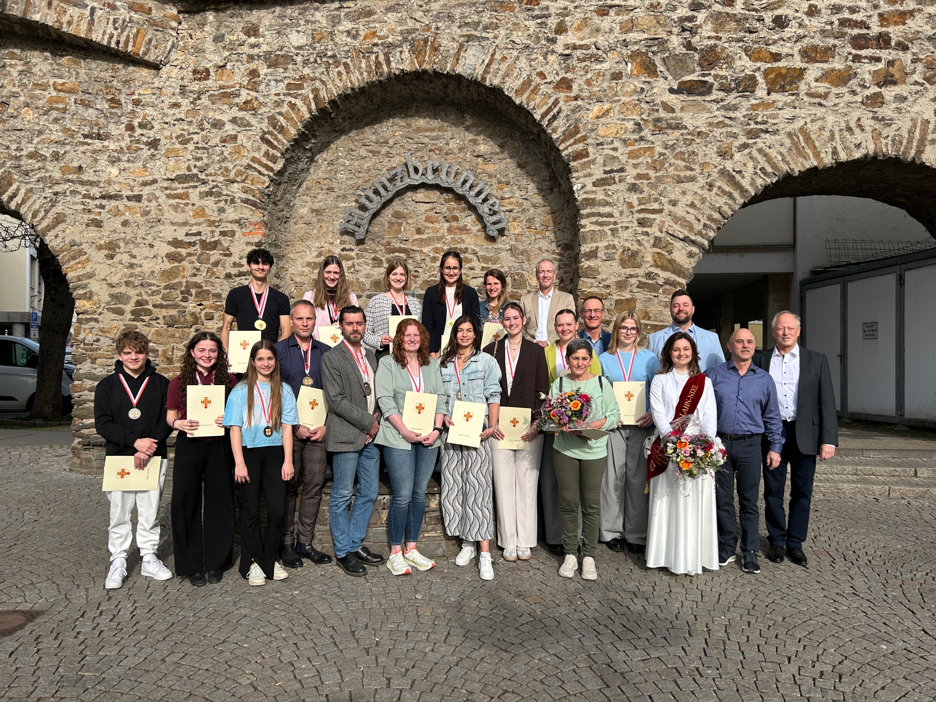 25 Personen stehen in zwei Reihen vor dem Münzbrunnen, einige tragen Medaillen um den Hals und Urkunden in den Händen.