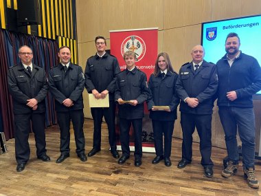 Eine Gruppe von Feuerwehrmännern und -frauen in Uniformen vor einer Holzwand und einem Bildschirm