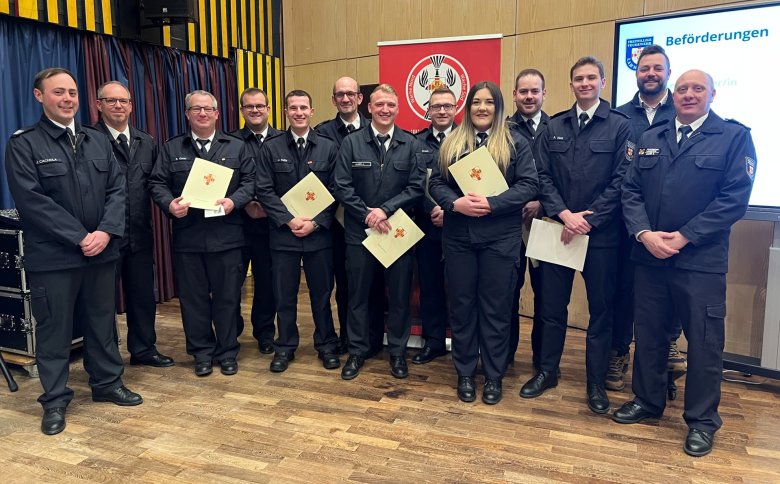 Eine Gruppe von Feuerwehrmännern und -frauen in Uniformen vor einer Holzwand und einem Bildschirm