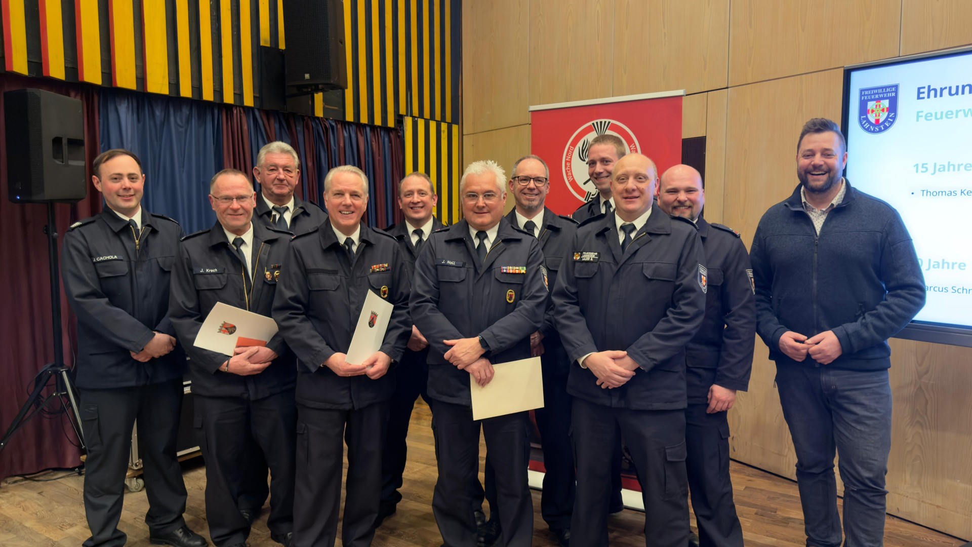 Eine Gruppe von Feuerwehrmännern in Uniformen vor einer Holzwand und einem Bildschirm