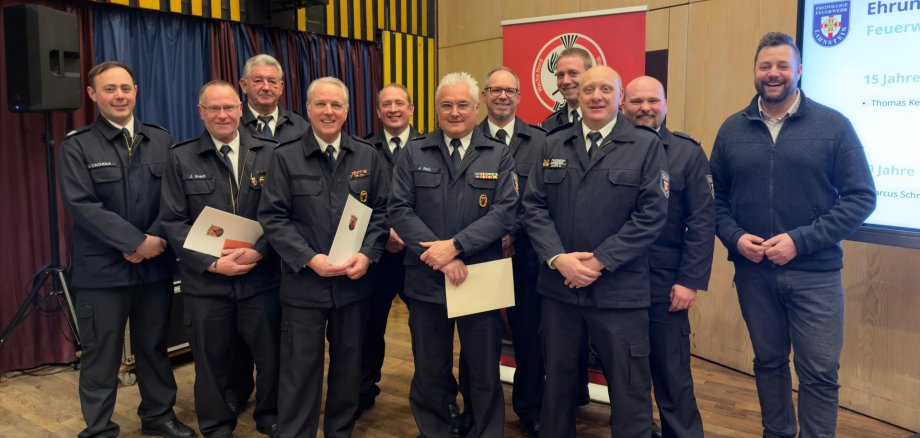 Eine Gruppe von Feuerwehrmännern in Uniformen vor einer Holzwand und einem Bildschirm