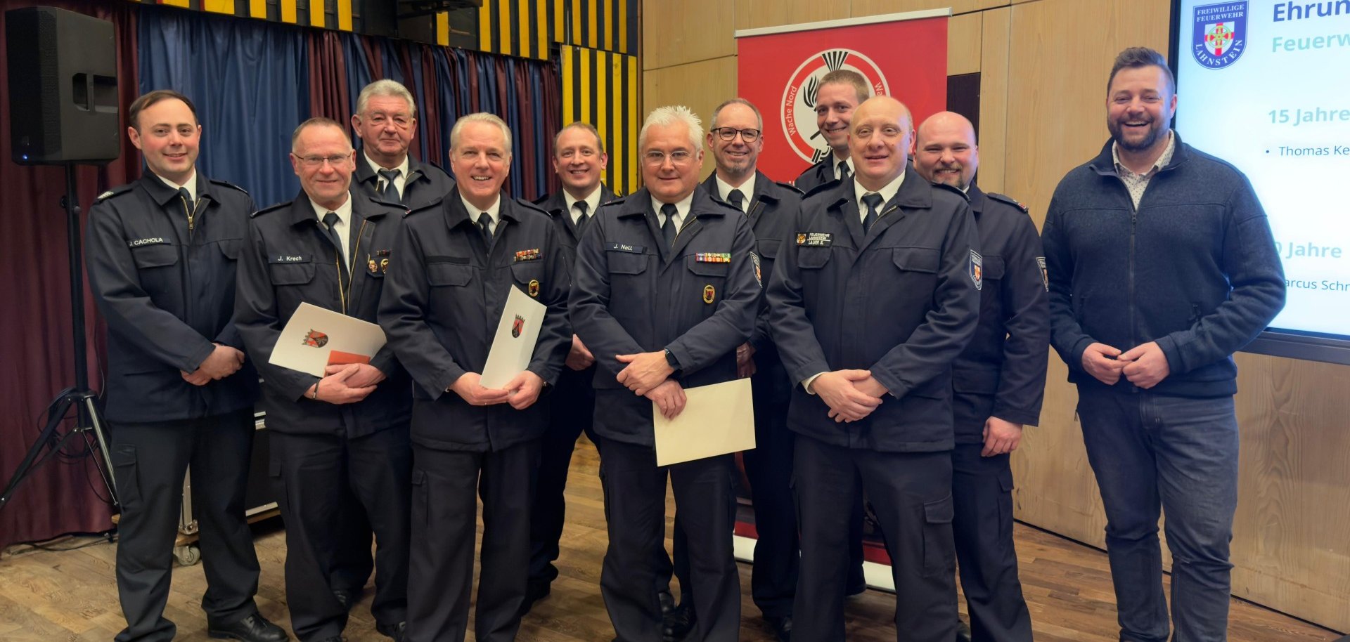 Eine Gruppe von Feuerwehrmännern in Uniformen vor einer Holzwand und einem Bildschirm