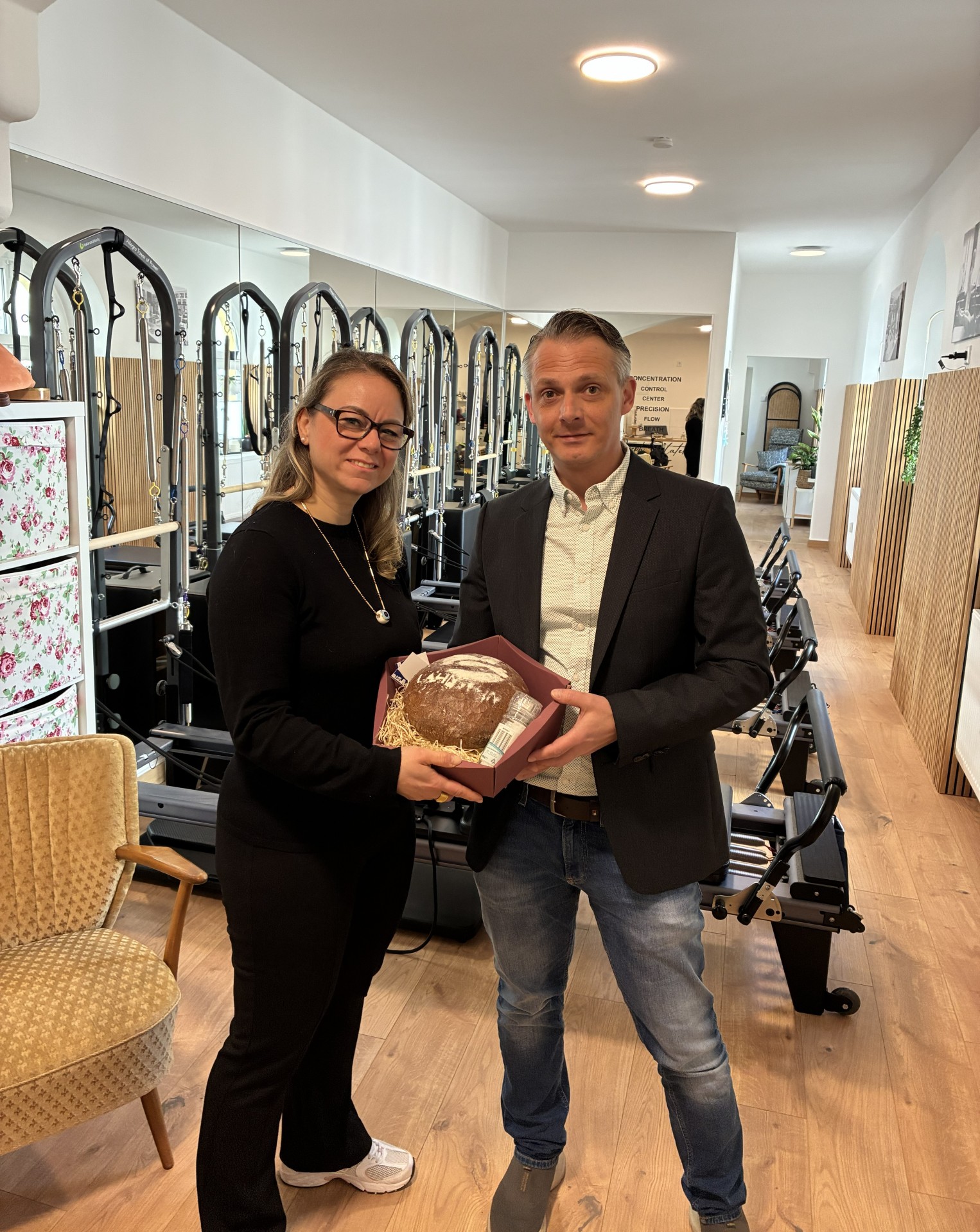 Die Inhaberin des Geschäfts "Just me" steht links neben Wirtschaftsförderer Christoph Noack in ihrem Studio. Im Hintergrund sind Fitnessgeräte zu sehen. Die zwei Personen schauen in die Kamera und halten gemeinsam einen Korb mit Brot und Salz.