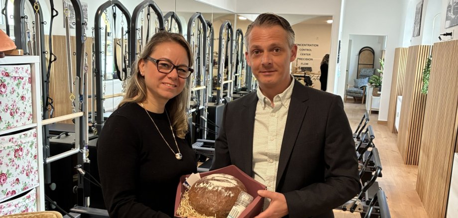 Die Inhaberin des Geschäfts "Just me" steht links neben Wirtschaftsförderer Christoph Noack in ihrem Studio. Im Hintergrund sind Fitnessgeräte zu sehen. Die zwei Personen schauen in die Kamera und halten gemeinsam einen Korb mit Brot und Salz.