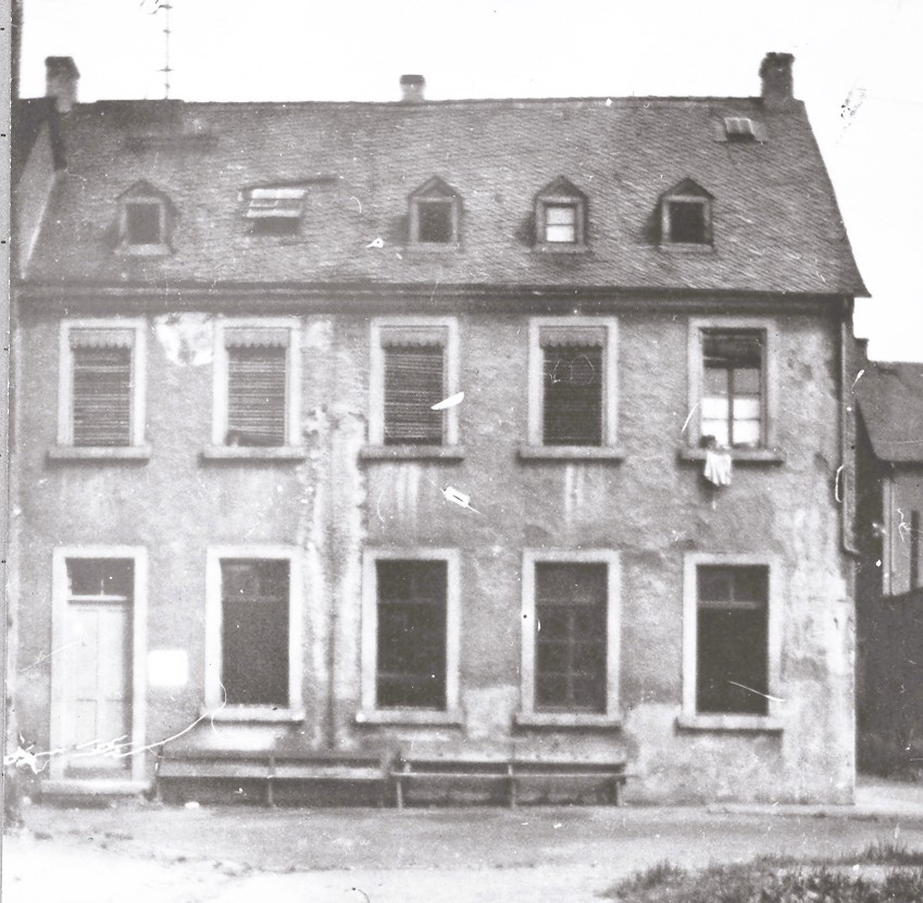 Schwarzweißfoto eines zweigeschossigen Hauses mit Dachgauben und einfacher Putzfassade. Vor dem Gebäude stehen Bänke; mehrere Fenster sind mit Rollläden versehen, eines ist geöffnet. Das Bild zeigt das Gebäude in älterem, teilweise verwittertem Zustand.