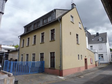 Vor 100 Jahren erhielt Niederlahnstein eine Jugendherberge Farbfoto eines Gebäudes aus schräger Straßenperspektive. Das zweigeschossige Haus mit ausgebautem Dachgeschoss ist gelb verputzt. Vor dem Gebäude befindet sich ein blauer Metallzaun, daneben eine schmale Straße und angrenzende Häuser
