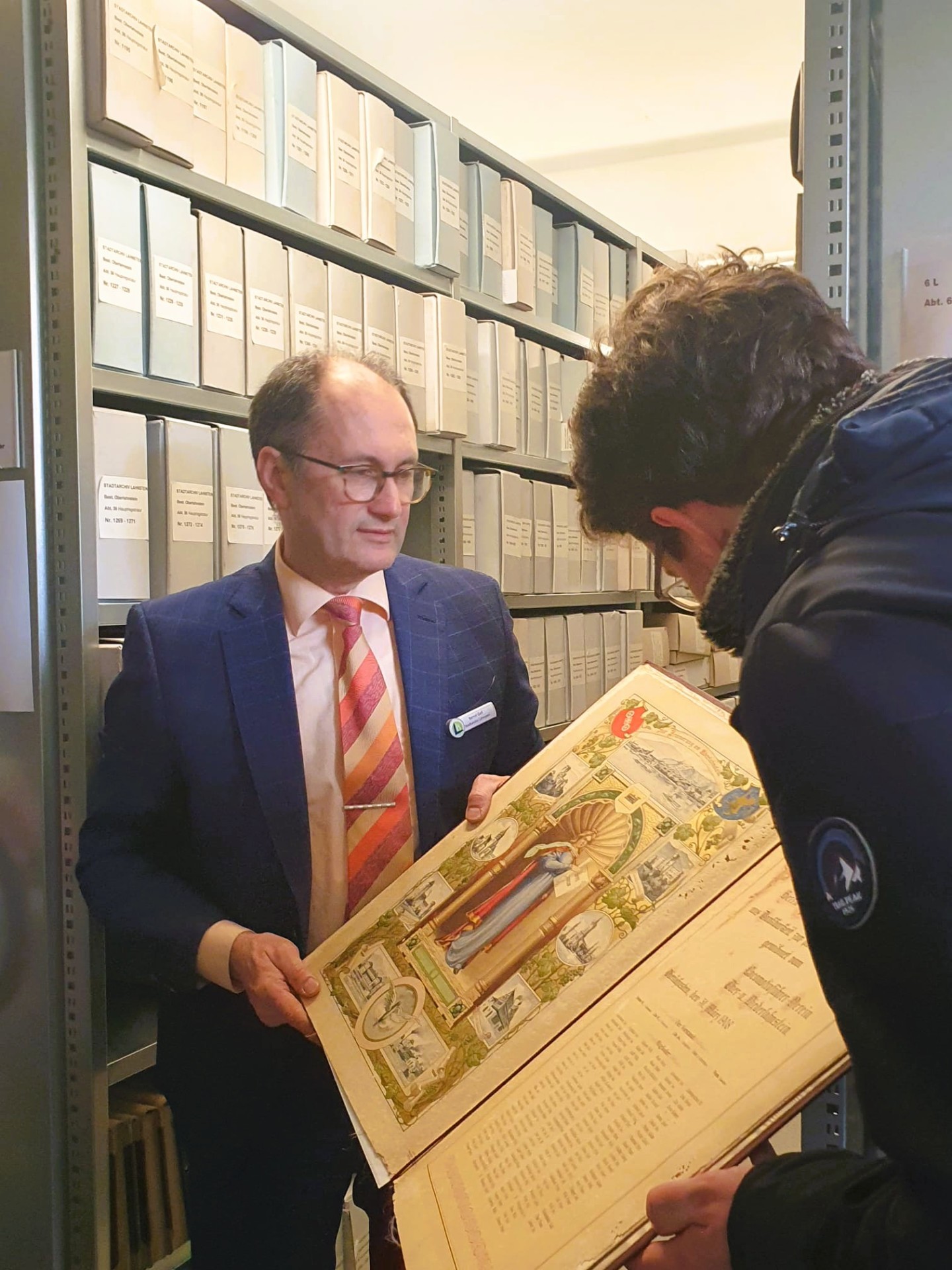 Zwei Männer stehen in einem Magazinraum des Stadtarchivs vor Regalen mit beschrifteten Archivkartons. Einer hält einen großen historischen Bildband geöffnet, während der andere sich darüber beugt und die reich illustrierte Seite betrachtet.