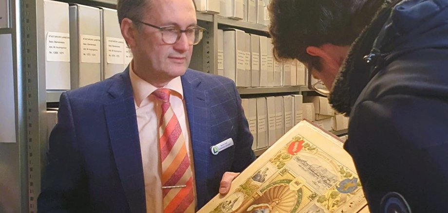 Zwei Männer stehen in einem Magazinraum des Stadtarchivs vor Regalen mit beschrifteten Archivkartons. Einer hält einen großen historischen Bildband geöffnet, während der andere sich darüber beugt und die reich illustrierte Seite betrachtet.