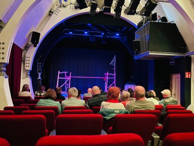 Publikum sitzt in roten Stuhlreihen eines kleinen Theatersaals und blickt auf eine Bühne mit einfacher Kulisse und blauem Bühnenlicht.“