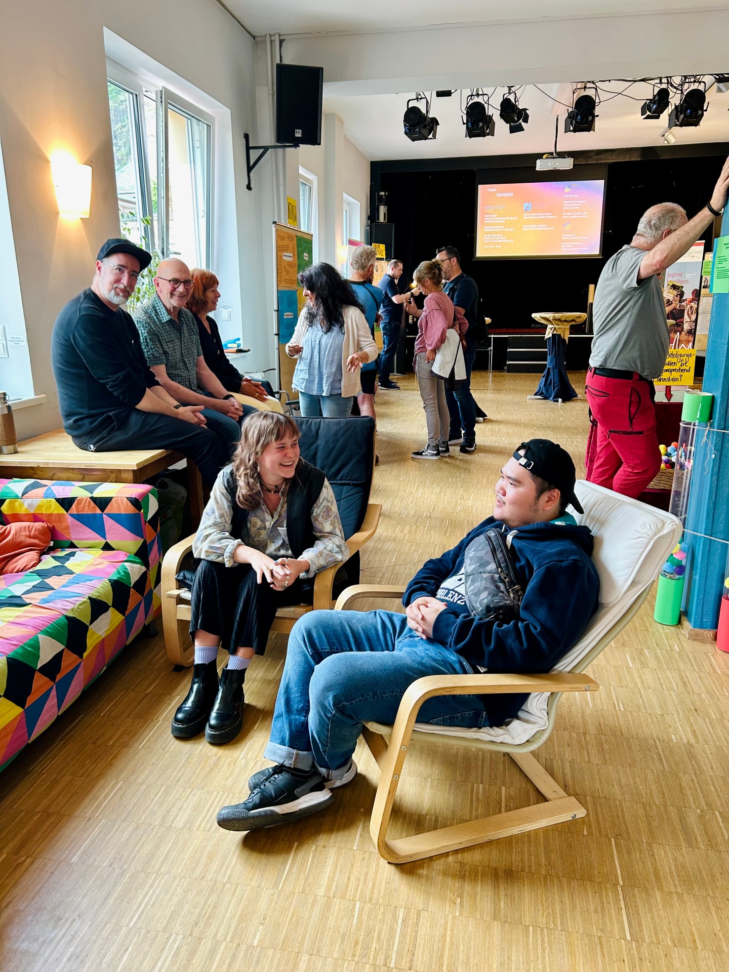 Mehrere Menschen unterhalten sich und sitzen in einem hellen Veranstaltungsraum mit Bühne im Hintergrund. Einige sitzen auf Sesseln und Bänken, andere stehen in kleinen Gruppen zusammen.