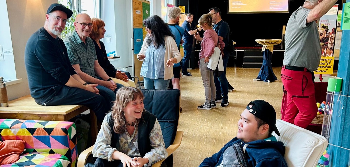 Mehrere Menschen unterhalten sich und sitzen in einem hellen Veranstaltungsraum mit Bühne im Hintergrund. Einige sitzen auf Sesseln und Bänken, andere stehen in kleinen Gruppen zusammen.