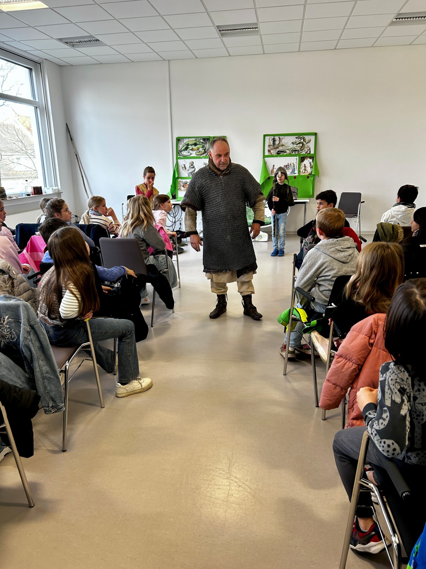 Ein Mann in mittelalterlicher Kleidung steht in der Mitte eines Stuhlkreises und spricht zu einer Gruppe von Kindern in einem hellen Raum. Die Kinder sitzen aufmerksam auf Stühlen um ihn herum. An der Wand hinter ihm hängen grüne Tafeln mit Bildern, und ein Kind steht daneben mit einem langen Stock oder Speer. Die Szene wirkt wie eine pädagogische Vorführung oder ein interaktiver Unterricht über das Mittelalter.
