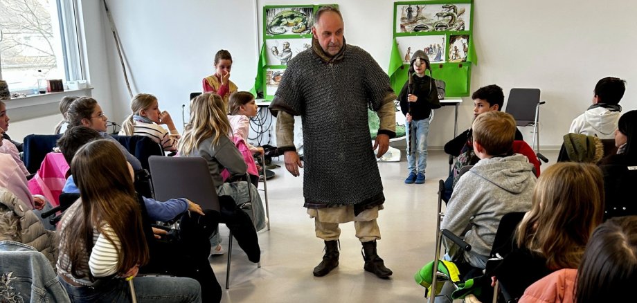 Lebendige Bücherei Ein Mann in mittelalterlicher Kleidung steht in der Mitte eines Stuhlkreises und spricht zu einer Gruppe von Kindern in einem hellen Raum. Die Kinder sitzen aufmerksam auf Stühlen um ihn herum. An der Wand hinter ihm hängen grüne Tafeln mit Bildern, und ein Kind steht daneben mit einem langen Stock oder Speer. Die Szene wirkt wie eine pädagogische Vorführung oder ein interaktiver Unterricht über das Mittelalter.