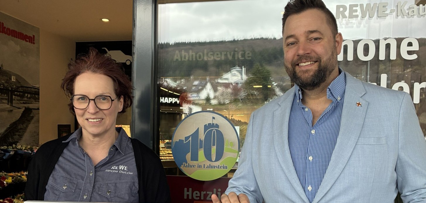 Simone Dietzler steht neben Oberbürgermeister Lennart Siefert vor der geöffneten Tür des des REWE Marktes Lahnstein. Sie halten eine Fotoleinwand in den Händen, auf der die Burg Lahneck zu sehen ist. Zwischen ihnen ist das runde Lahnstein-Label mit einer großen 10 auf die Scheibe des Eingangs geklebt.