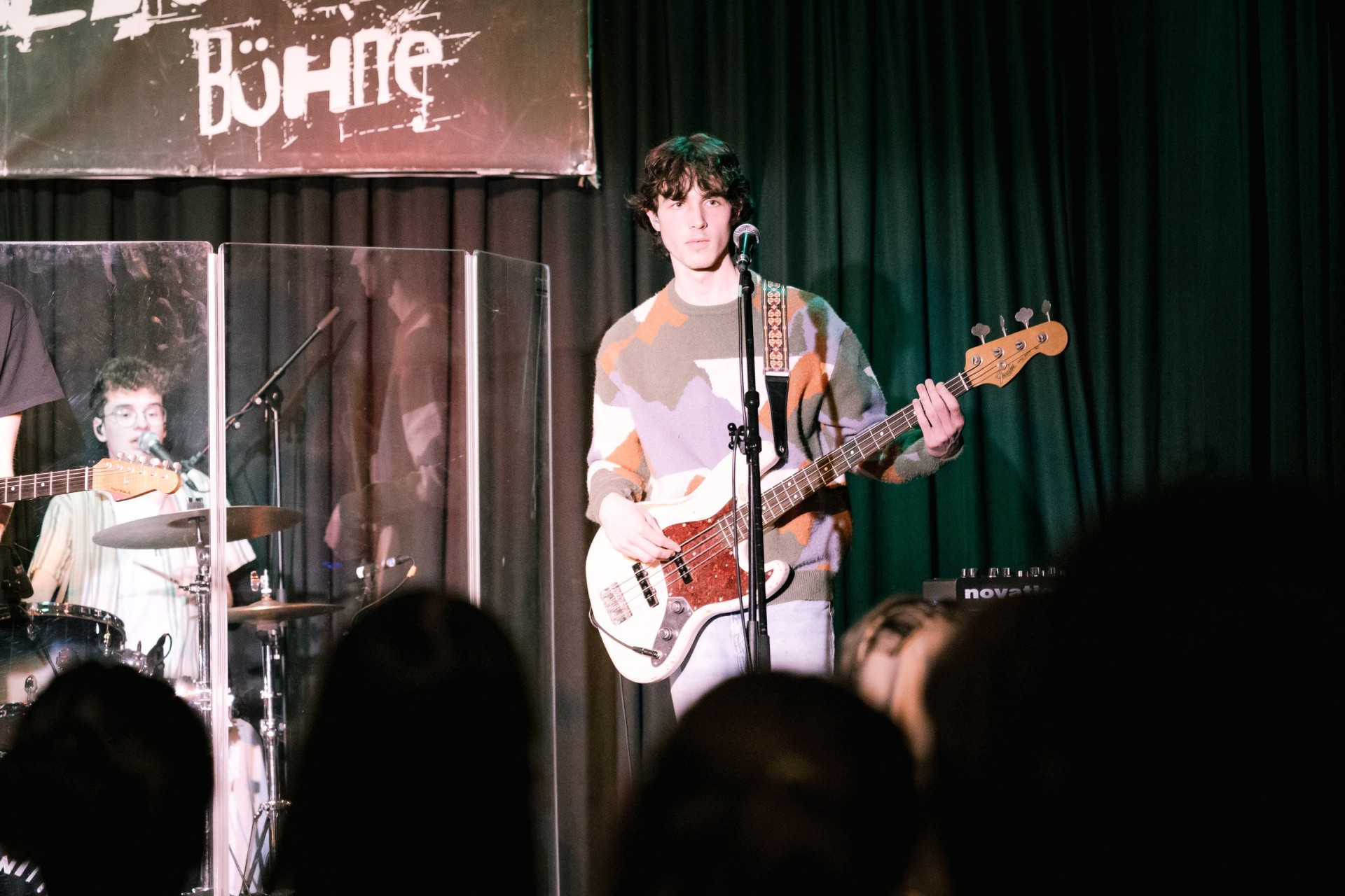 Ein Musiker spielt E-Bass und singt auf einer Bühne, im Hintergrund Schlagzeuger, im Vordergrund Publikum.