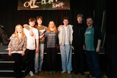Volles Haus beim Konzert von Herbstwind im Jugendkulturzentrum Eine Gruppe von sechs jungen Menschen und einem erwachsenen Mann steht gemeinsam auf einer Bühne im Jugendkulturzentrum vor einem schwarzen Vorhang. Einige legen sich freundschaftlich die Arme um die Schultern. Über ihnen hängt ein Schild mit der Aufschrift „Bühne“. Alle schauen in die Kamera und lächeln.