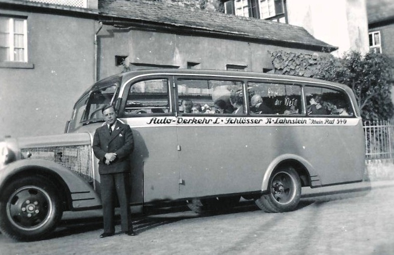Schwarzweißfoto: Ein Mann im Anzug steht vor einem historischen Linienbus mit der Aufschrift eines Verkehrsunternehmens.