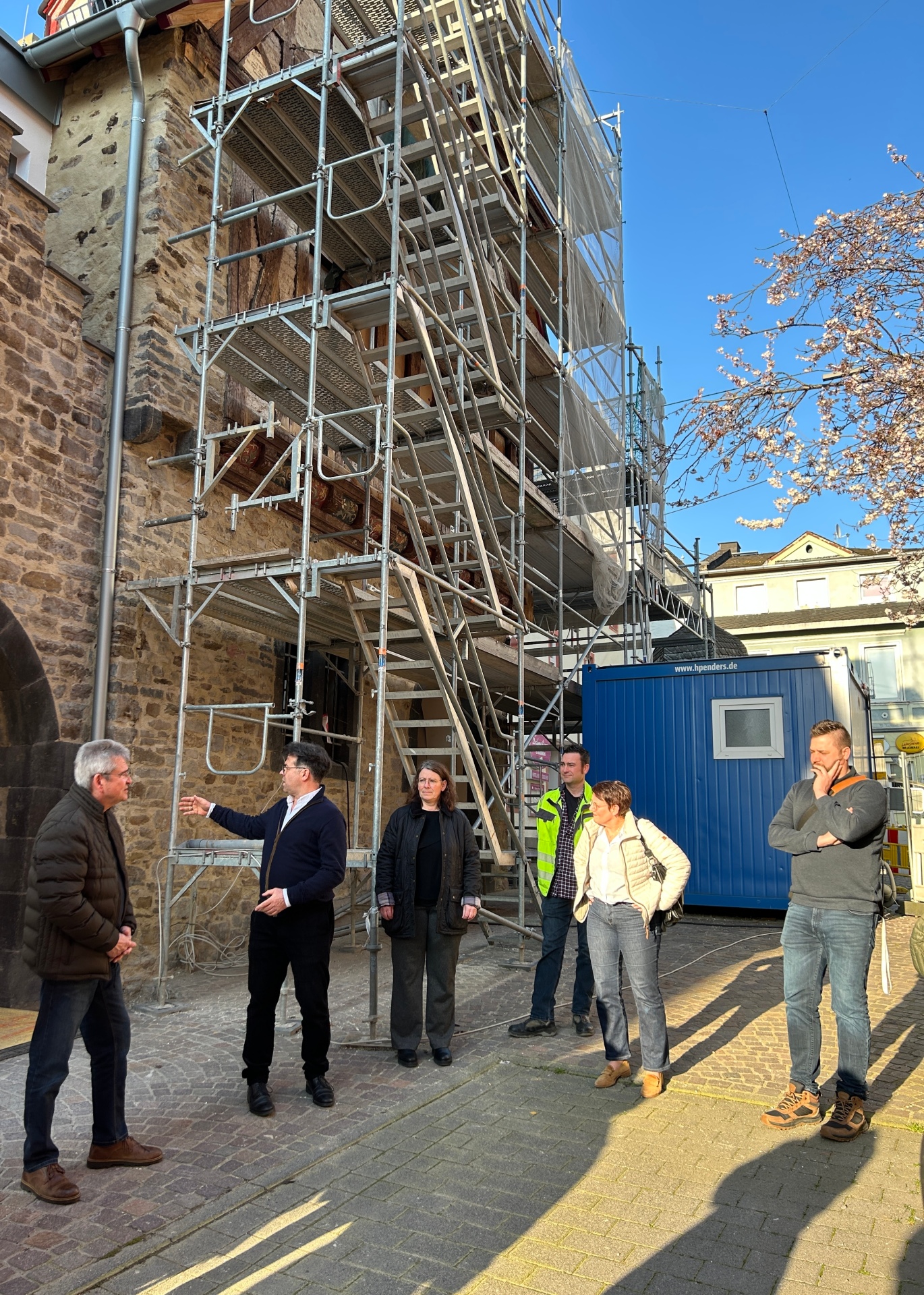 Mehrere Personen stehen bei einer Baustellenbegehung vor dem Alten Rathaus und betrachten die Arbeiten. An der historischen Steinfassade ist ein großes Gerüst mit Treppen aufgebaut. Eine Person erklärt gestikulierend Details zur Baustelle, während die anderen aufmerksam zuhören. Im Hintergrund steht ein blauer Baucontainer, daneben sind weitere Gebäude zu sehen. Sonniges Wetter mit klarem Himmel und blühenden Ästen am rechten Bildrand.